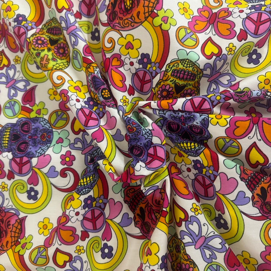 A close-up of a colorful patterned fabric with a tie-dye or knotted center, featuring a retro psychedelic design with skulls, peace signs, butterflies, and hearts in pink, yellow, purple, orange, and green on a cream background.