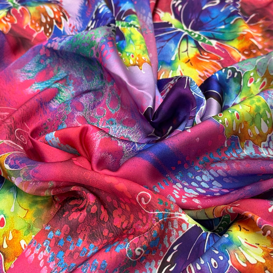 A close-up of a vibrant, multicolored silk scarf with a butterfly print and watercolor-like splashes of pink, blue, purple, green, and yellow.