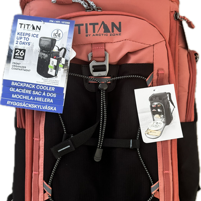 A Titan by Arctic Zone backpack in terracotta pink and black, shown with its included cooler accessory packaging and an informational hangtag against a plain white background.
