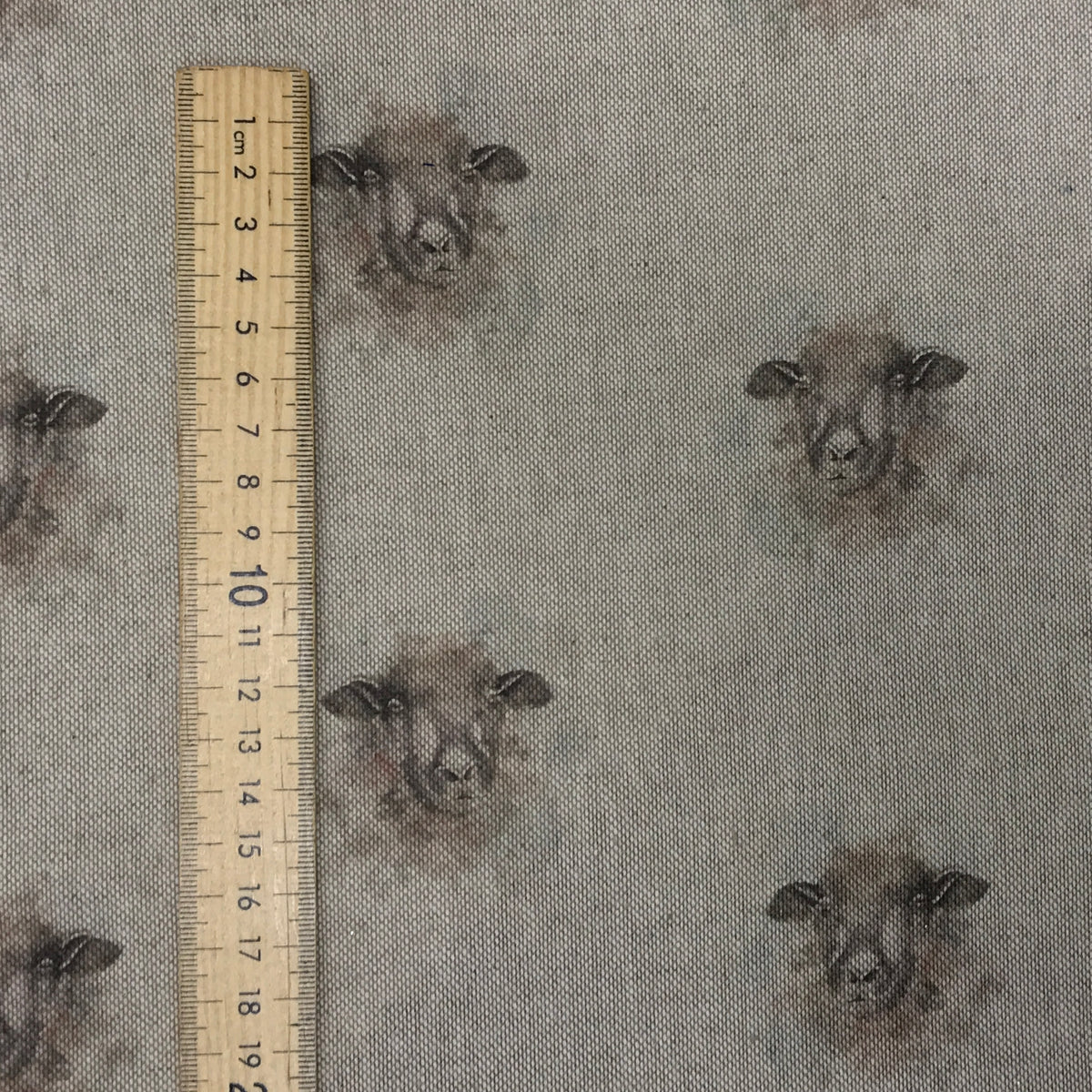 A close-up of a woven fabric with a repeating print of sheep faces in dark brown and gray tones on a light beige background, shown next to a wooden ruler for scale.