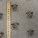 A close-up of a woven fabric with a repeating print of sheep faces in dark brown and gray tones on a light beige background, shown next to a wooden ruler for scale.