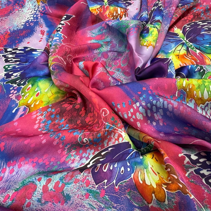 A close-up of a draped silk scarf featuring a vibrant, multicolored butterfly and floral print with pink, purple, blue, yellow, and green tones.