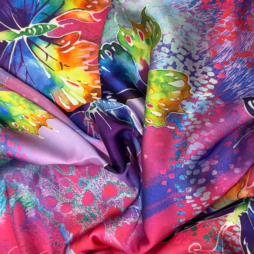 A close-up of a draped silk scarf featuring a vibrant, multicolored butterfly print with watercolor-like splashes of pink, purple, blue, green, and yellow.