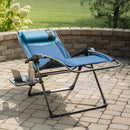 A blue and black outdoor folding lounge chair with a side tray table, set on a paved patio with a brick wall and green lawn in the background. A laptop and a bottle are placed on the tray table.