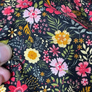 A close-up of a black fabric with a dense, colorful floral print featuring pink, yellow, and white flowers with green and gold leaves. A person's finger is visible on the left side holding the fabric.