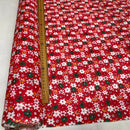 Red fabric with a dense white, green, and yellow floral print, shown partially rolled with a wooden ruler running vertically to indicate scale.