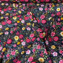A close-up of a floral fabric with a dense pattern of pink, red, yellow, and white flowers with green leaves on a black background, draped with soft folds.