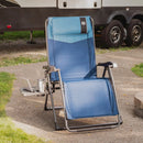 A blue and light blue outdoor folding lounge chair with a headrest, black armrests, and a side tray table holding a laptop and cup, set on a paved surface next to grass with an RV in the background.