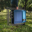 A person outdoors in a grassy park area is holding a folded camping chair with a black metal frame and blue seat cushions.