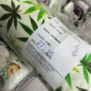 A close-up of a roll of printed cotton poplin fabric with a green cannabis leaf pattern on an ivory background, wrapped in clear plastic with a white paper label showing details.