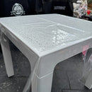A clear plastic waterproof table cover draped over a white plastic table, with water droplets on the transparent surface. The background shows a dark gray tiled floor and black branded tarps.
