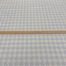 A close-up of a light blue and white houndstooth patterned fabric, with a wooden ruler laid horizontally across the center for scale.