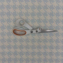 A pair of scissors with white plastic handles and orange accents, resting on a light blue and white houndstooth patterned fabric background.